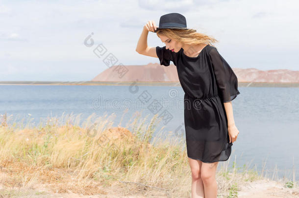 美丽的年轻金发女人，穿着黑色的连衣裙，戴着一顶浅黑色的帽子，在炎热的夏天，风吹着她的头发