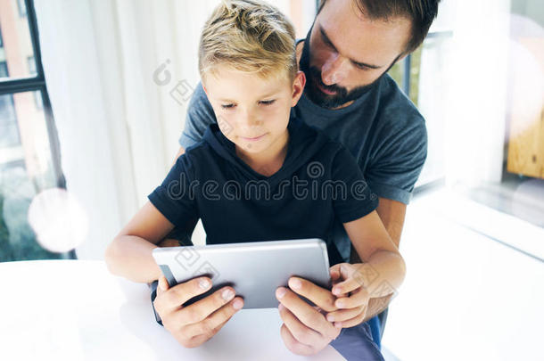 父亲和他的小儿子一起在移动电脑上玩耍，休息在室内。胡须男和小男孩在阳光明媚的时候使用平板电脑