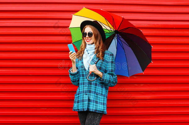 <strong>时尚</strong>漂亮的年轻微笑的女人，带着<strong>五</strong>颜六色的雨伞，使用智能手机，穿着一件红色的<strong>黑</strong>色帽子外套
