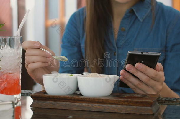 黑发女孩坐在咖啡馆外面吃<strong>冰淇淋</strong>时使用智能<strong>手机</strong>。 4k，