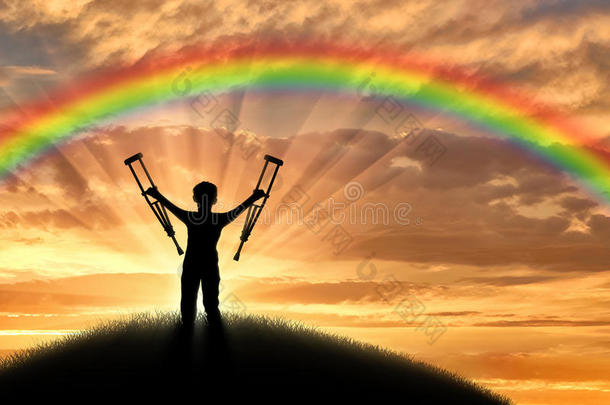 残疾儿童拄着拐杖站在彩虹背景上的山上日落。