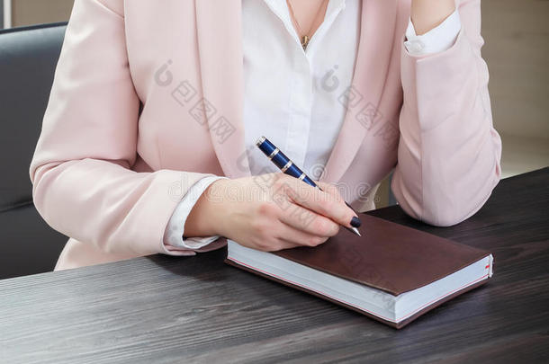 迷人的黑发女人穿着米色西装，坐在办公室的桌子上，拿着笔记本。 特写镜头