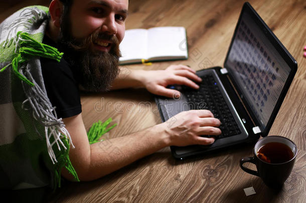蓄着胡子的英俊男子躺在地板上，裹着格子和工作笔记本电脑