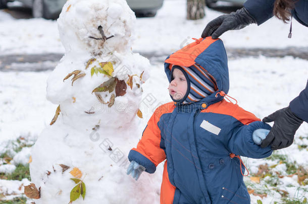 迷人的小男孩玩第一场雪。 他微笑着看着雪人。 厚厚的蓝色橙色连体裤，明亮的条纹