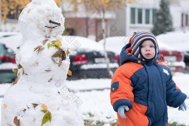 迷人的小男孩玩第一场雪。 他微笑着看着雪人。 厚厚的蓝色橙色连体裤，明亮的条纹