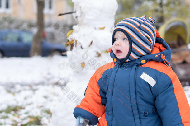 迷人的小男孩玩第一场雪。 他微笑着看着雪人。 厚厚的蓝色橙色连体裤，明亮的条纹