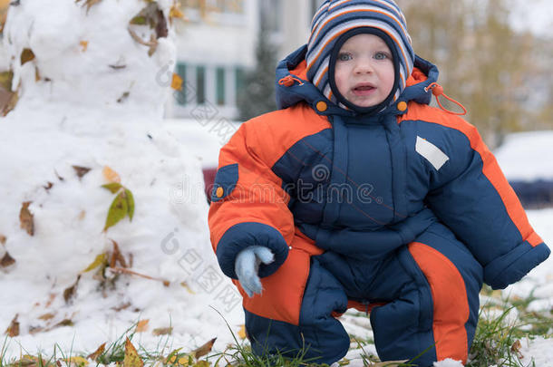 迷人的小男孩玩第一场雪。 他微笑着看着雪人。 厚厚的蓝色橙色连体裤，明亮的条纹