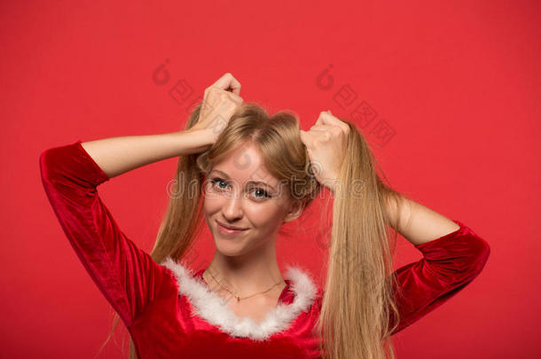 穿着圣诞老人服装的美丽的年轻女人玩着一缕丝质的长发，看着红色背景上的相机