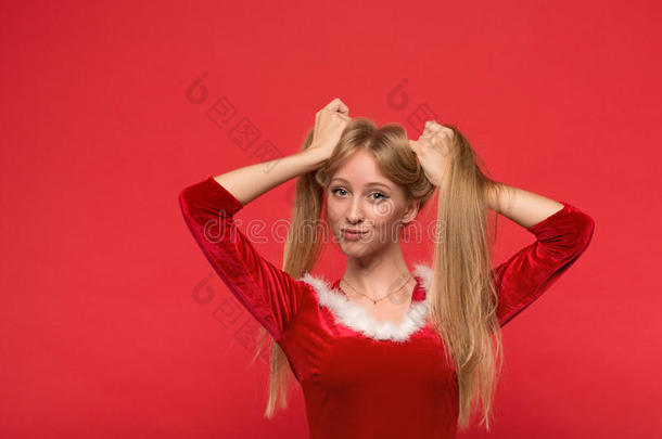 穿着圣诞老人服装的美丽的年轻女人玩着<strong>一缕</strong>丝质的长发，看着红色背景上的相机