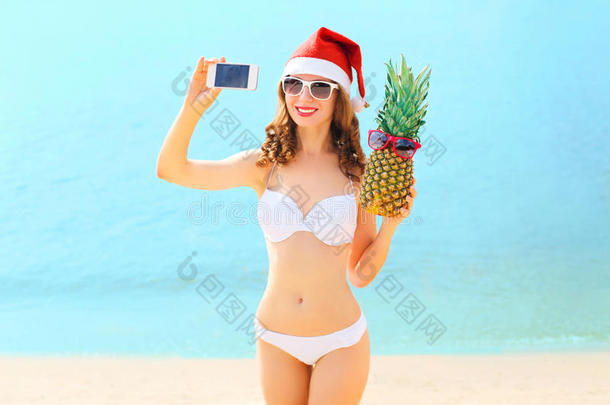 圣诞微笑的年轻女子戴着红圣诞帽，戴着菠萝在海边的智能手机上自拍