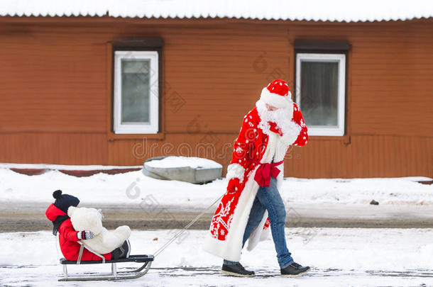 爸爸穿着圣诞老人的西装，他的小儿子在冬天的雪下骑着雪橇，在乡村街道上