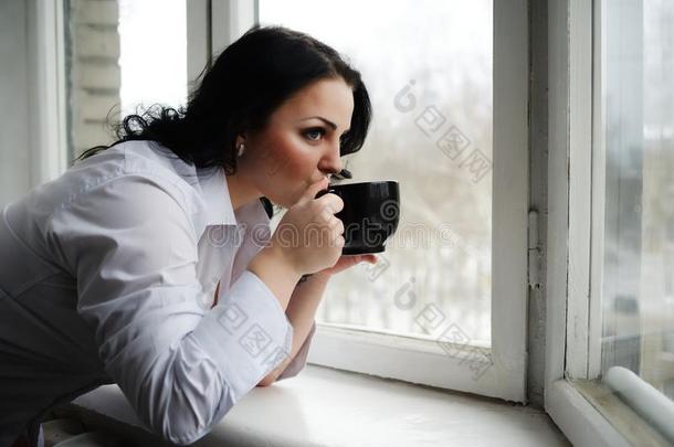 漂亮的女孩看着窗外，拿着一个杯子。 冬天的早晨。