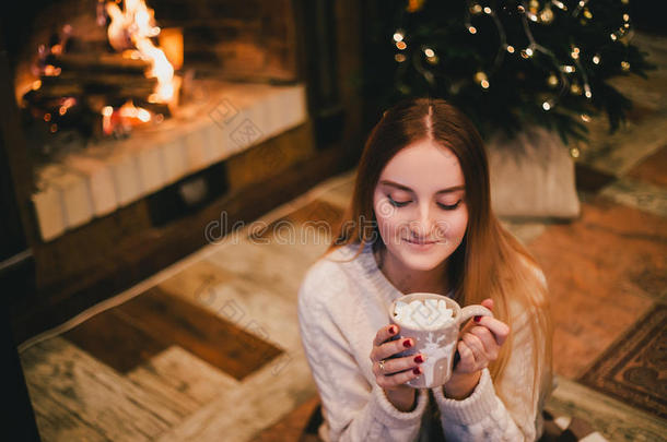 美丽的年轻女人坐在圣诞树下的壁炉旁，喝着棉花糖的可可