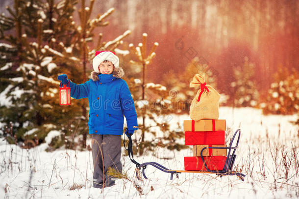 戴着圣诞老人帽子的可爱小男孩在雪林里拿着一只木制雪橇和礼物