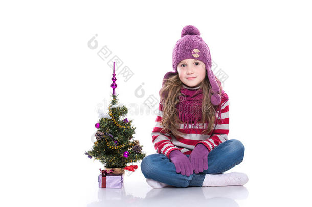 可爱微笑的小女孩戴着紫色针织围巾和帽子，坐在圣诞树附近，礼物被隔离在白色的背景上。