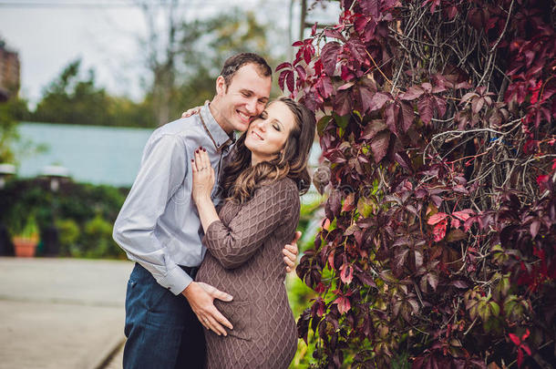 一个怀孕的年轻女人和她的丈夫。一个幸福的家庭站在红色的秋篱前，抱着肚子。孕妇放松