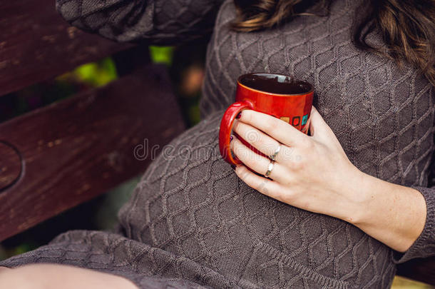 孕妇的肚子坐在秋千上，拿着一杯茶。 孕妇在公园放松。