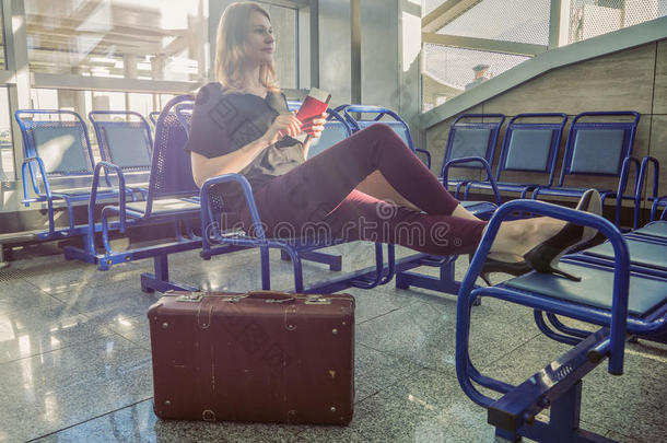 漂亮的女孩坐在大厅里，等待航班。 梦想度假。 带复古行李箱的旅程。