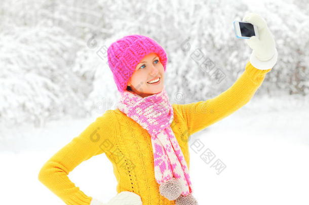 时尚冬季快乐微笑的年轻女人在智能手机上拍摄自己的肖像，戴着五颜六色的针织帽子，在雪地树上