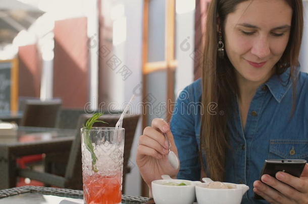 黑发女孩坐在咖啡馆外面吃<strong>冰淇淋</strong>时使用智能<strong>手机</strong>。 4k，