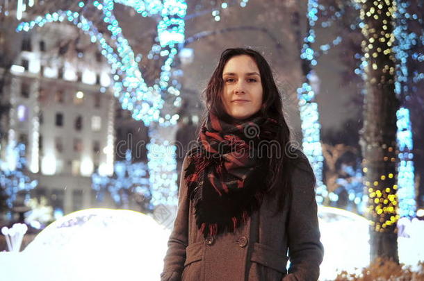 迷人的女人在下雪的<strong>圣诞夜</strong>微笑着看着公园树木前装饰着闪闪发光的灯光的相机
