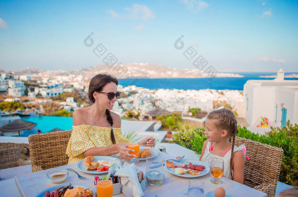 一家人在户外咖啡馆吃早餐，在Mykonos镇有令人惊叹的景色。 可爱的女孩和妈妈喝新鲜果汁