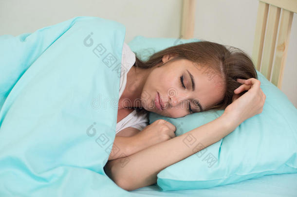 美丽的年轻女人睡在家里的床上，在蓝色的羽绒被下，手臂靠近她的脸