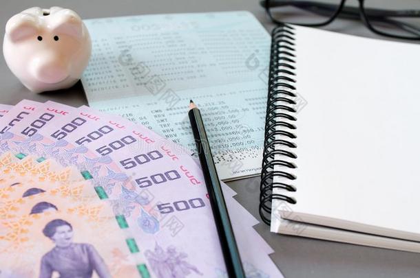 灰色背景上的空白笔记本、铅笔、储蓄账户存折、眼镜、泰国货币和储蓄罐