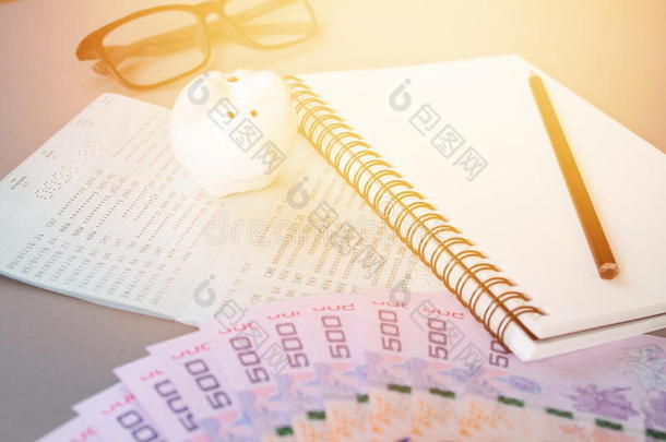 灰色背景上的空白笔记本、铅笔、储蓄账户存折、眼镜、泰国货币和储蓄罐