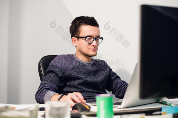 忙着工作。坐在工作场所，戴着眼镜，坐在笔记本电脑前的年轻帅哥