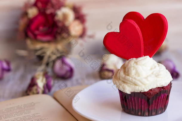 在一本打开的书的书页上，一个白色盘子上装饰着两颗红心的纸杯蛋糕。