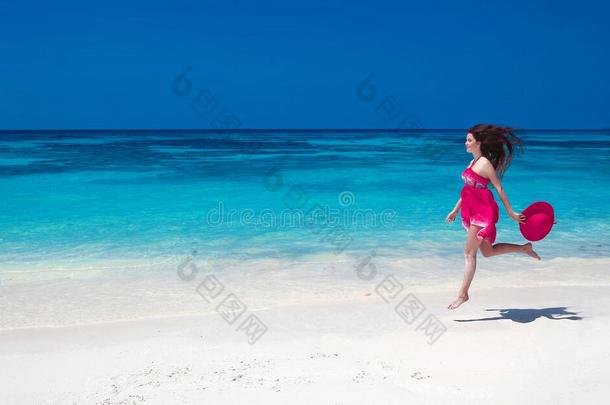 美丽的自由年轻女子在异国情调的海洋上跳跃，快乐的黑发女孩穿着粉红色的连衣裙在热带海滩上奔跑，沙滩上有白沙