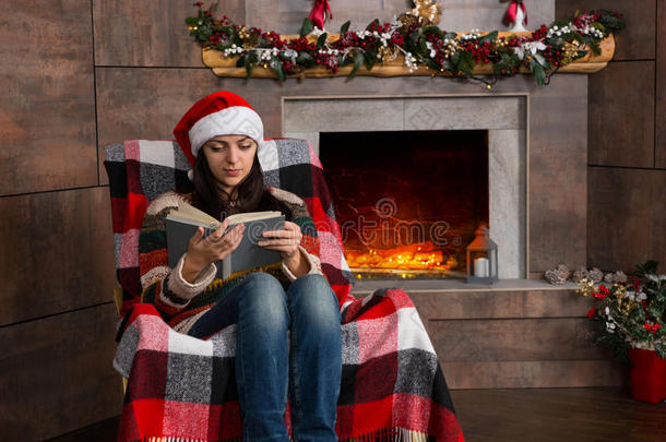 迷人的集中妇女戴着有趣的圣诞帽阅读，而坐在圣诞树附近的摇椅上
