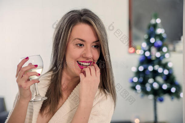 女孩在圣诞树旁喝酒，思考。 美丽的女人坐在圣诞树旁边，享受着一杯酒