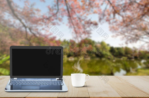 电脑笔记本电脑，黑色屏幕和热咖啡杯在木制桌面上模糊的湖泊和樱花树背景