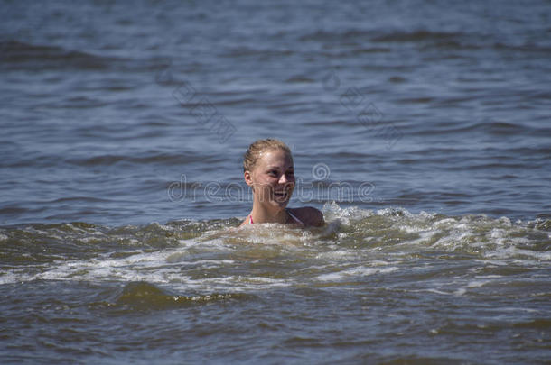 金发女孩笑着，喜欢在海水里游泳。 女孩在海水的背景下浸入水中直到他们的脖子。 是