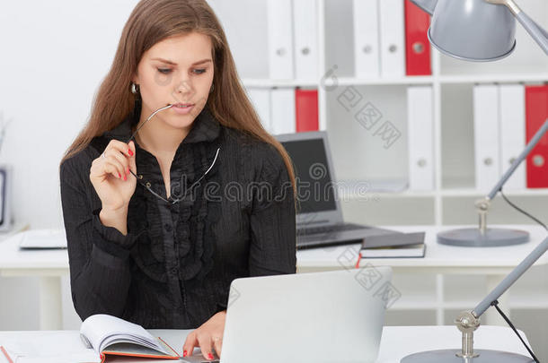 漂亮的年轻秘书拿着眼镜看着坐在办公室的笔记本电脑。