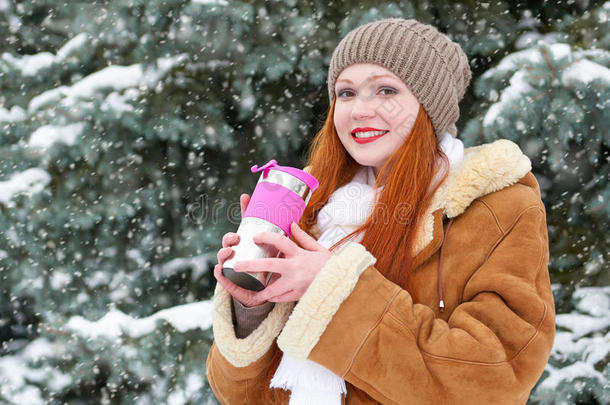 美丽的女人喝一杯热饮，在冬天的户外保暖，森林里的雪杉树，红色的长发，穿着羊皮