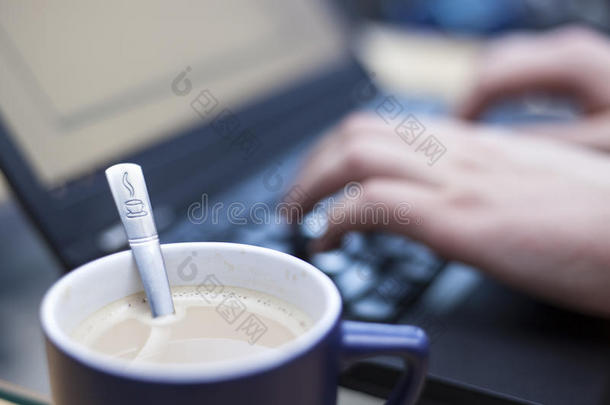 一杯咖啡，<strong>上面</strong>印有咖啡标志的茶匙，焦点是一个人在后台的<strong>笔记本</strong>电脑上工作