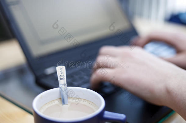 一杯咖啡，上面印有咖啡标志的茶匙，焦点是一个人在后台的笔记本电脑上工作