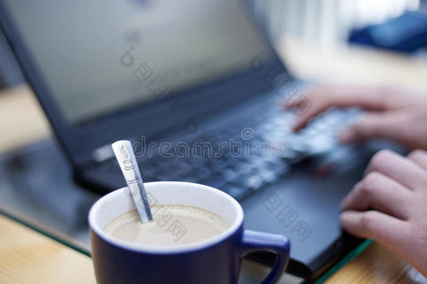 一杯咖啡，上面印有咖啡标志的茶匙，焦点是一个人在后台的笔记本电脑上工作