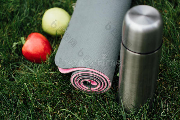 在草地上的公园里，有吸引人的灰色粉<strong>红色滚动</strong>瑜伽或健身垫，里面有苹果和热水瓶。
