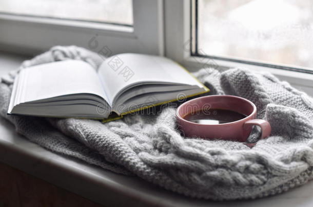 舒适的家庭静物：一杯热咖啡和打开的书与温暖的格子在窗台上抵御外面的雪景