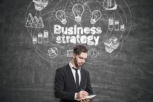 商人拿着<strong>笔记本</strong>站在黑板附近，<strong>上面</strong>画着商业战略。