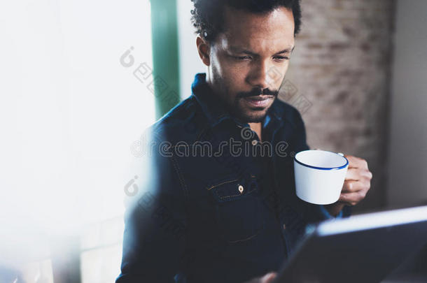 近景的年轻胡须非洲男子使用平板电脑，同时手持<strong>白色</strong>陶<strong>瓷杯</strong>在现代合作