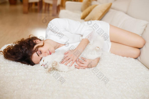 美丽的女人，卷曲的头发，穿着白色的丝绸长袍，一大早就和白色的毛茸茸的猫坐在沙发上玩
