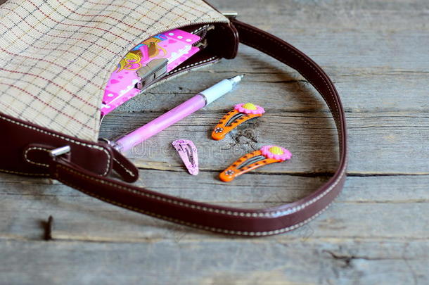 木制桌子上的儿童手提包和配件。 小手提包，笔记本，粉红色钢笔，明亮的发夹给女孩