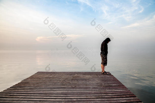 沮丧的年轻人穿着黑色连帽衫站在木桥上，伸向大海，俯视着大海，沉思着自杀