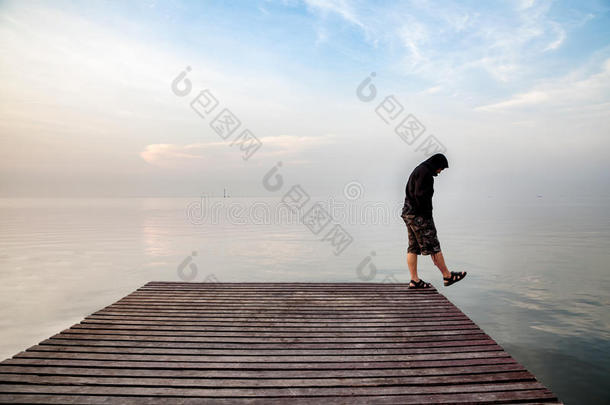 沮丧的年轻人穿着黑<strong>色</strong>连帽衫站在木桥上，伸向<strong>大海</strong>，俯视着<strong>大海</strong>，沉思着自杀
