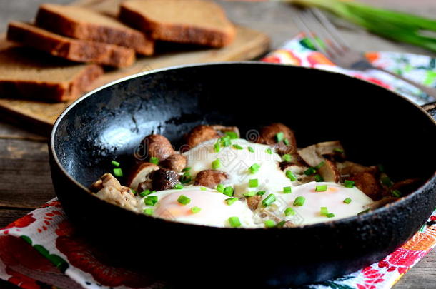 煎鸡蛋与蘑菇在煎锅，棕色面包片，新鲜洋葱，叉子在木桌上
